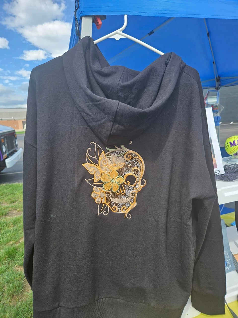Gray hoodie with a gold sugar-skull and floral embroidery on the back, shown at a craft fair